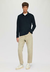 Polo a maniche lunghe blu navy con taschino sul petto, abbinato a jeans beige a gamba affusolata e sneakers bianco sporco con suola testurizzata.