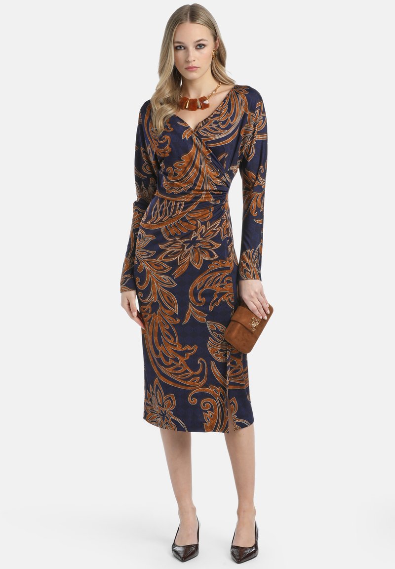 Middellange mouwen marineblauwe jurk met oranje bloemenpatronen, een getailleerde silhoeët en een wikkeldesign. Het model houdt een bruine clutch vast en draagt hakken.