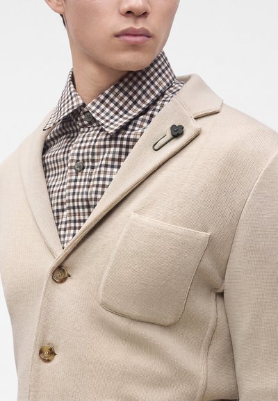 Homme portant un blazer beige texturé avec une épingle noire en forme de fleur et une chemise à carreaux marron et blanc boutonnée.