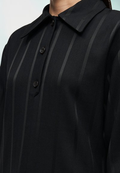 Camicia nera con scollo a collo, tre bottoni e tessuto strutturato con sottili righe verticali per un interesse visivo.