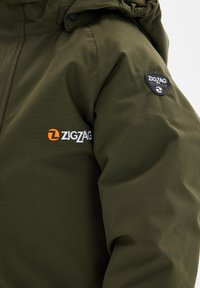 Olijfgroene jas met een gladde textuur, capuchon en geborduurd "ZIGZAG" logo in oranje en wit op de linkermouw. Zwart patchlogo geïntegreerd.