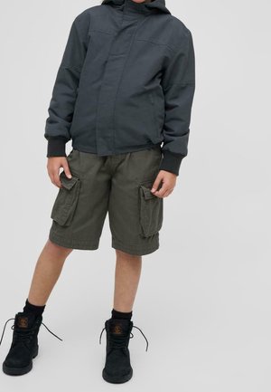 Niño que lleva una chaqueta oscura con capucha, pantalones cortos cargo verdes con grandes bolsillos, calcetines negros y botas negras hasta el tobillo, de pie sobre un fondo liso.