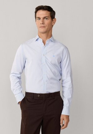 Hombre con camisa a rayas azul claro de manga larga y pantalones marrón oscuro, de pie con una mano en el bolsillo.