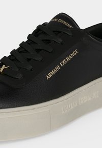 Baskets en cuir noir avec des lacets noirs, lettrage doré "Armani Exchange" sur le côté et la languette, et une semelle épaisse blanc cassé.