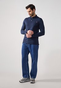 Polo de manga larga en color navy con un bolsillo en el pecho, combinado con jeans de corte relajado en azul y zapatillas deportivas grises con un diseño estampado.