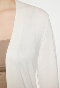 Lichte beige cardigan met lange mouwen, open voorkant en geribbelde textuur. De stof lijkt zacht en iets doorzichtig.