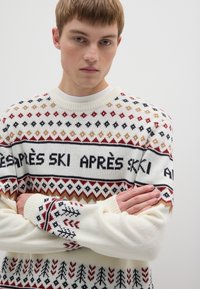 Strickpullover in Creme mit mehrfarbigem Muster, mit dem Schriftzug "APRÈS SKI" und geometrischen Formen, mit geripptem Hals- und Bündchenabschluss.