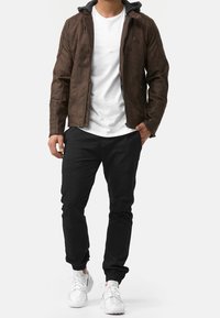 Brun kunstlæderjakke med hætte og grå indvendig side, tætsiddende design, lynlås foran og ribbet detaljering. Båret med sorte bukser og hvide sneakers.