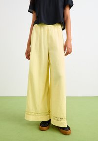 Light yellow wide-leg pants made of textured fabric, featuring a decorative lace trim at the hem. Paired with black footwear.