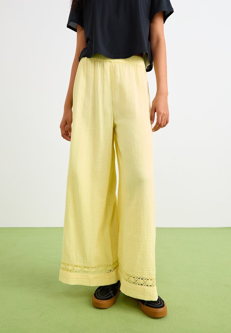 Light yellow wide-leg pants made of textured fabric, featuring a decorative lace trim at the hem. Paired with black footwear.