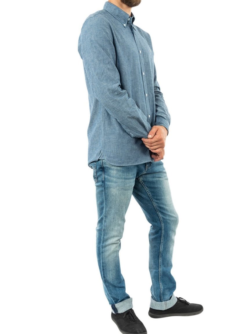 Camicia di denim azzurro chiaro con colletto button-down, maniche lunghe e una sottile texture, abbinata a jeans blu sbiaditi e scarpe nere.