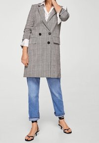 Cappotto a doppio petto a quadri in tessuto grigio e nero, caratterizzato da due grandi bottoni neri, abbinato a jeans arrotolati e sandali neri.