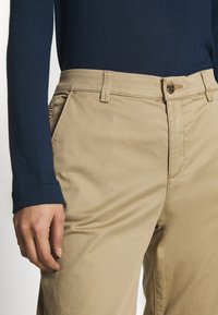 Pantaloni di cotone beige con una texture liscia, dotati di chiusura anteriore con bottone e tasche laterali. Indossati con un top a maniche lunghe blu scuro.