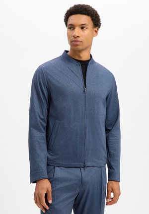 Mann trägt eine blaue Jacke mit Reißverschluss und passende Hose, steht mit entspannten Händen und schaut leicht zur Seite.