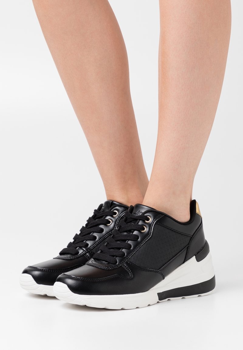 Svarta syntetiska sneakers med en strukturerad panel, rund tå, vit sula och gulddetalj vid hälen. Skosnören genom metallögla.