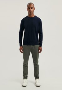 Maglione a maniche lunghe di colore blu navy realizzato in tessuto testurizzato, abbinato a pantaloni slim-fit color verde oliva e sneakers bianche. Design semplice e moderno.