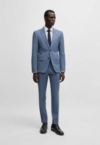 Lichtblauw op maat gemaakt pak met een subtiel ruitpatroon, enkelvoudig gestructureerd jasje, met notch revers, twee voordo pockets, gecombineerd met een witte shirt en een marineblauwe stropdas.