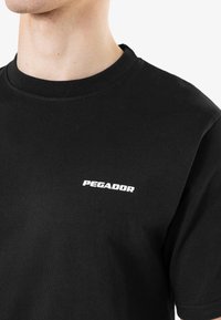 Melnā kokvilnas t-kreklu ar apaļu kakla izgriezumu un baltu "PEGADOR" logo, kas iespiests kreisajā krūtīs. Gluda tekstūra, ikdienišķs dizains.