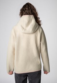 Vrouw met een gebroken-witte fleecejas met capuchon, van achteren gezien, met bruin krullend haar en donkergrijze broek tegen een effen achtergrond.