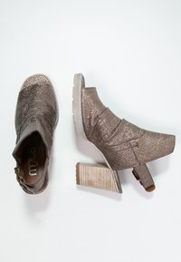 Metallinen pronssinvärinen nilkkaboots, jossa on tekstuuri pinnassa, varustettu lohkare korkokengällä ja säädettävällä remmillä soljella.