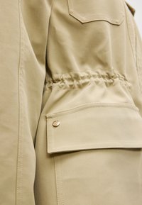 Μπεζ λειτουργικό μπουφάν με απαλό ύφασμα, ελαστική μέση, πλαϊνή τσέπη με καπάκι και κουμπί, και τετράγωνη τσέπη στο πίσω μέρος.