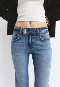 Svetlo modri denim kavbojke z visokim pasom, dvema sprednjima žepoma in gumbom za zapiranje. Teksturen material z rahlim zbledelostjo in kontrastnim šivanjem.
