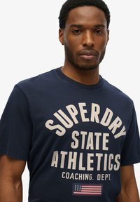 Camiseta de algodón azul marino con gran texto blanco "SUPERDRY STATE ATHLETICS", con un pequeño gráfico de la bandera americana en la parte inferior.