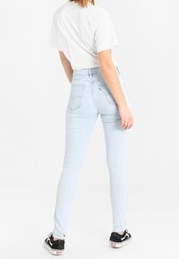 Pantalones vaqueros ajustados de talle alto en azul claro con una textura suave, que presentan dos bolsillos traseros y un dobladillo deshilachado, combinados con zapatillas negras y blancas.