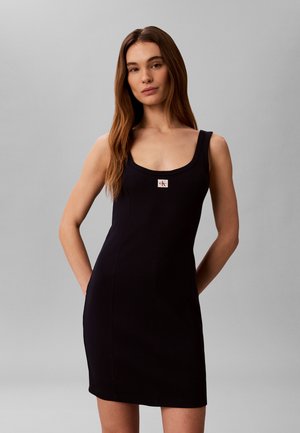 Γυναίκα με μακριά καστανά μαλλιά, φορώντας μαύρο μίνι φόρεμα χωρίς μανίκια με μικρό λογότυπο Calvin Klein στο στήθος, στέκεται μπροστά σε απλό γκρι φόντο.