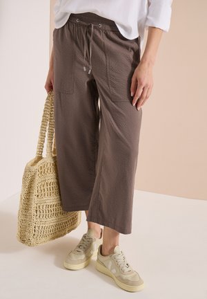Casual outfit met cropped bruine broek met voorzakken, beige sneakers en een grote beige geweven tote bag die een persoon aan zijn zijde vasthoudt.