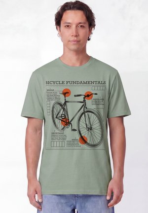 Homme portant un t-shirt vert clair avec un schéma de vélo noir et des parties étiquetées intitulé Fondamentaux du vélo.