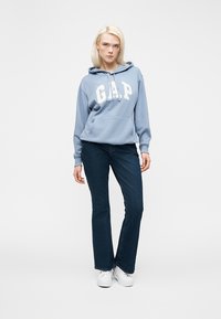 Jeune femme portant un sweat à capuche GAP bleu clair, un jean bleu foncé et des baskets blanches, debout devant un fond blanc uni.