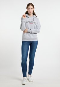 Felpa grigia con tasca a marsupio frontale e cordini bianchi, caratterizzata da testo "DREIMASTER MARITIM" in nero e rosso. Indossata con jeans blu e scarpe da ginnastica bianche.