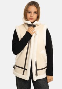 Jeune femme ajustant la boucle d'un gilet en peau retournée beige porté sur un haut à manches longues noir et un jean foncé, devant un fond uni.