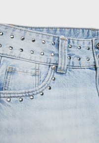 Jean en denim bleu clair en gros plan montrant la taille, les passants de ceinture, la poche avant, et des clous en métal argenté décorant la taille et les bords de la poche.