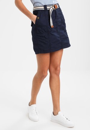 Kvinde iført en navy nederdel med lommer og et stribet bælte, kombineret med hvide sneakers og en blåstribet skjorte.