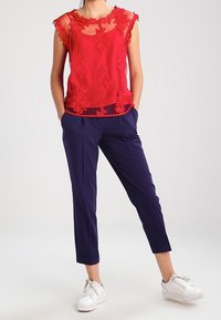 Rød blondebluse med blomsterbroderi og flæseærmer, parret med marineblå cropped bukser og hvide sneakers. Glat stof, tætsiddende design.