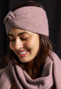 Pletený růžový čelenka s vykrouceným vzorem, texturovaným povrchem a odpovídajícím růžovým šátkem přehozeným kolem krku, umístěný na tmavém pozadí.