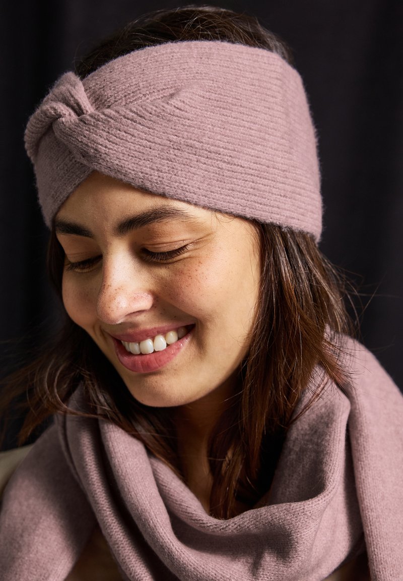 Pletený růžový čelenka s vykrouceným vzorem, texturovaným povrchem a odpovídajícím růžovým šátkem přehozeným kolem krku, umístěný na tmavém pozadí.