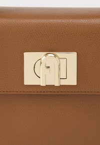 Brūna teksturēta ādas soma ar zelta toņā taisnstūra formu aizdari, kurā iespiests zīmola nosaukums "Furla". Gluda virsma un elegants dizains.