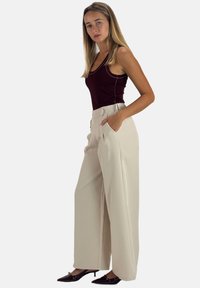 Pantalon beige à jambes larges avec poches avant, design ajusté et texture lisse, associé à un débardeur bordeaux.