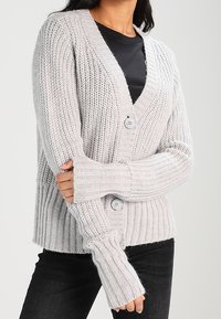 Cardigan lavorato a maglia grigio chiaro con scollo a V, due bottoni, texture a coste e maniche lunghe. Abbinato a una maglietta nera e pantaloni scuri.