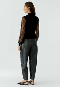 Femme de profil portant un haut en dentelle noir à manches longues, un pantalon en simili cuir noir et des escarpins à talon aiguille noirs sur fond uni.
