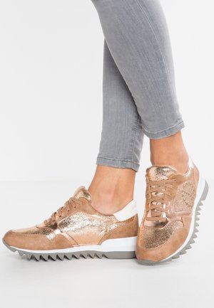 Voeten met glanzende rosegouden sneakers met witte en grijze zolen, gecombineerd met lichtgrijze nauwsluitende broek tegen een effen witte achtergrond.