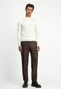 Cremfarbener gestrickter Pullover mit hohem Kragen, kombiniert mit braunen, maßgeschneiderten Hosen. Das Outfit zeichnet sich durch eine strukturierte Optik und einen figurschmeichelnden Schnitt aus.