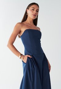 Námornícka modrá šaty bez ramienok s priliehavým živôtikom, plisovanou sukňou a vreckami. Vyrobené z hladkej látky, s čistým dizajnom a minimálnymi detailmi.