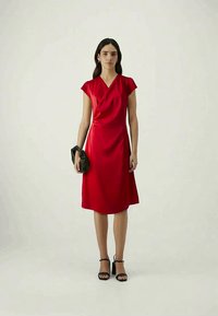 Vestido de cetim vermelho com mangas curtas, design cruzado, silhueta até ao joelho. Modelo a segurar uma bolsa preta; a usar sandálias de salto pretas.