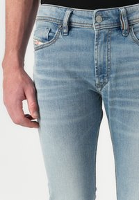 Jeans din denim albastru deschis cu o croială slim, având cusături în contrast și o ușoară decolorare. Include buzunare frontale și închidere cu un nasture metalic.