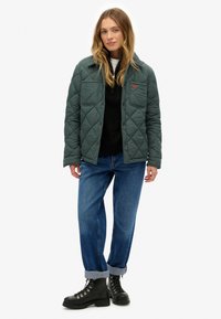 Chaqueta acolchada verde con cierre de botones y dos bolsillos en el pecho, combinada con pantalones vaqueros de mezclilla azul con puños enrollados y botas negras de cordones.