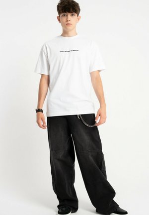 Młody mężczyzna w białej koszulce z napisem "TAKE A MOMENT TO BREATHE", czarnych szerokich spodniach z łańcuchem i czarnych butach.